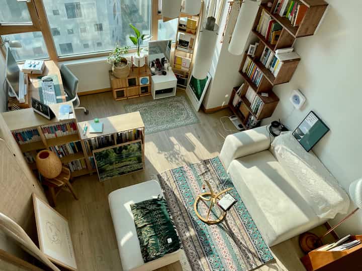 ウッドトーンとホワイトを基調としたコンパクトなリビング兼ホームオフィス、書棚とソファが配置された自然な雰囲気の空間