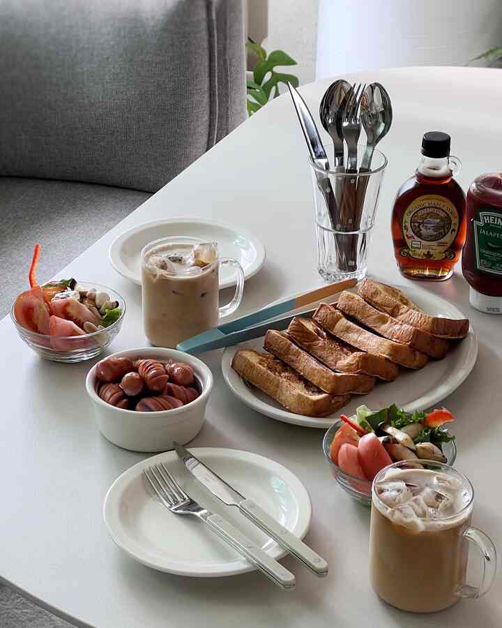 White-toned dining table featuring breakfast set with toast, sausages, salads, and iced coffee in a cafe-style setting