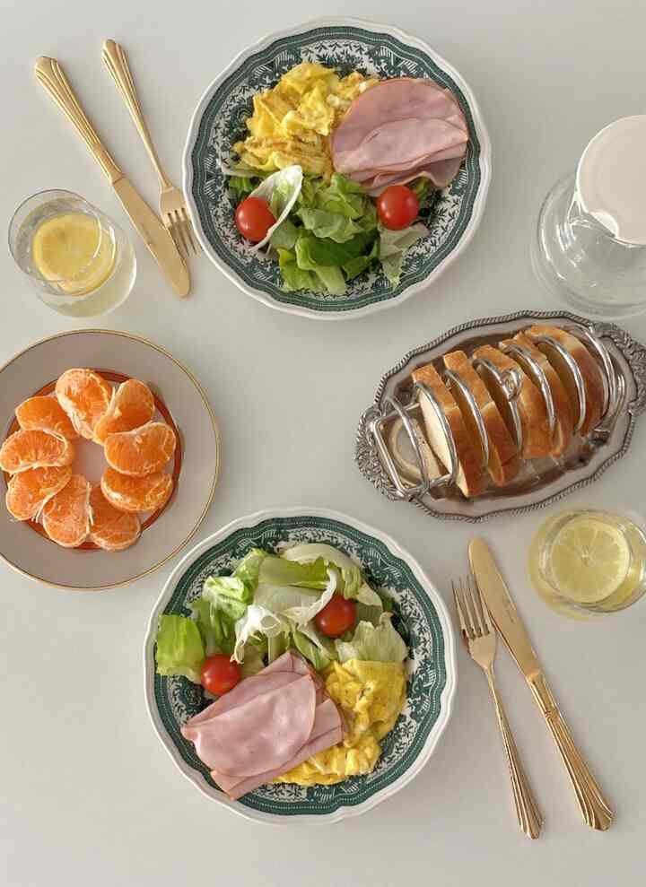 Simple and clean plating on a white table featuring two vintage green patterned plates, gold cutlery, and a silver tray with bread slices