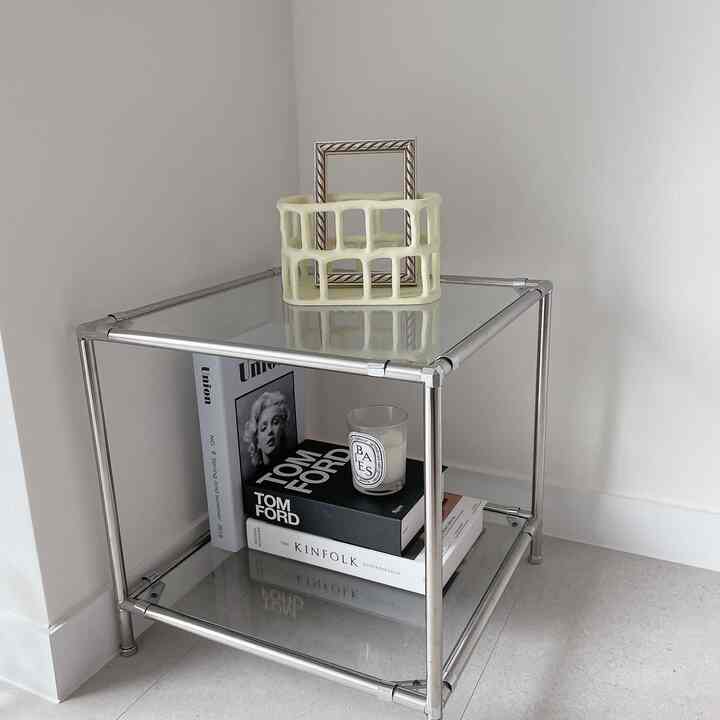 白い壁とシルバーメタルフレームのモジュール式ナイトテーブルにインテリアブックとキャンドルが配置されたミニマルな空間