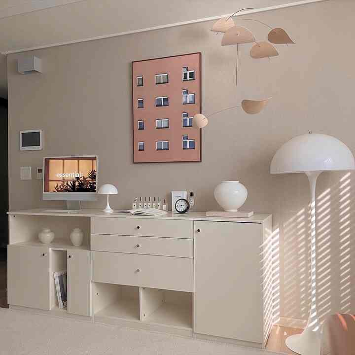 Natural beige-toned living space featuring white dresser, table and floor lamps, artistic wall art and mobile creating a cozy atmosphere