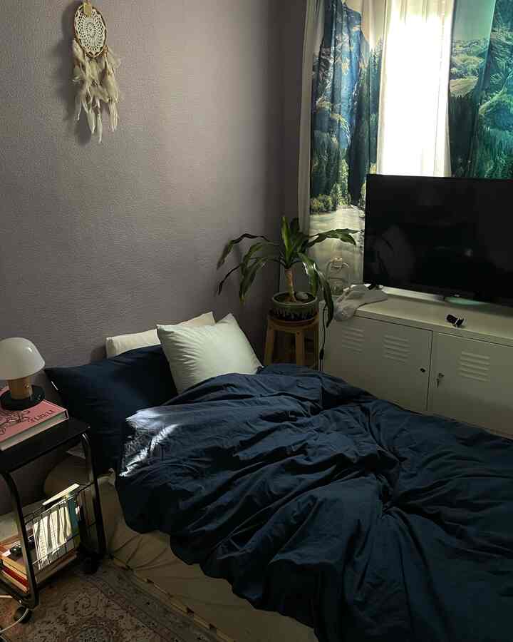 Cozy bedroom featuring navy duvet cover and white pillows, wooden stool with plant, and dreamcatcher on muted wall