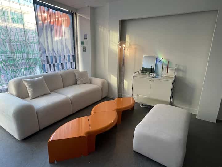 White-toned living room featuring a central orange coffee table, spacious sofa, floor lamp, and cabinet in a simple modern space