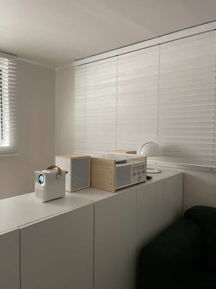 Living room in white and wood tones, featuring blinds, audio equipment, and minimal furniture for a cozy atmosphere