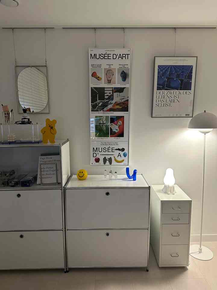 グレーの壁と白のモジュール家具、ミッドセンチュリーモダン風の大きなポスターとフロアランプが配置された在宅勤務用インテリア