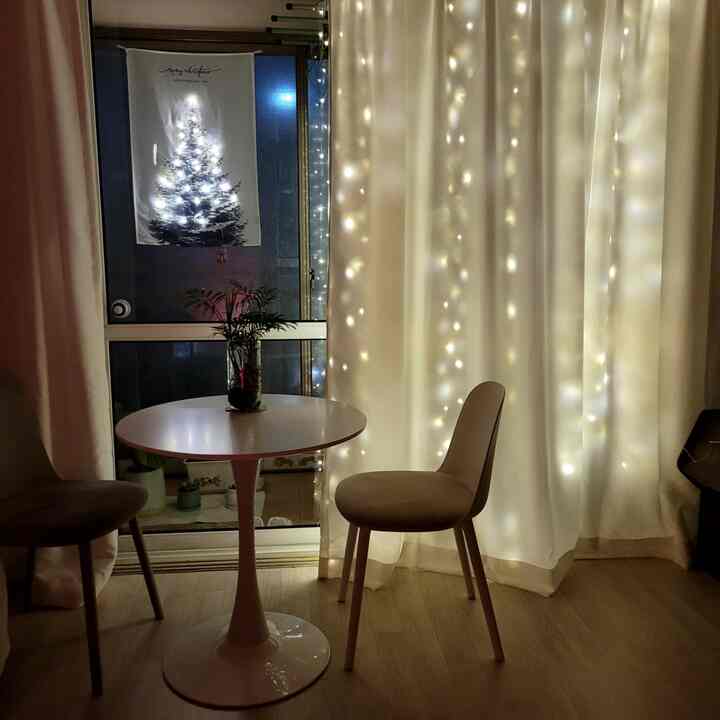 白いカーテンと暖かいストリングライトがあるダイニングルーム、小さな丸いテーブルと椅子が配置されたアットホームな空間
