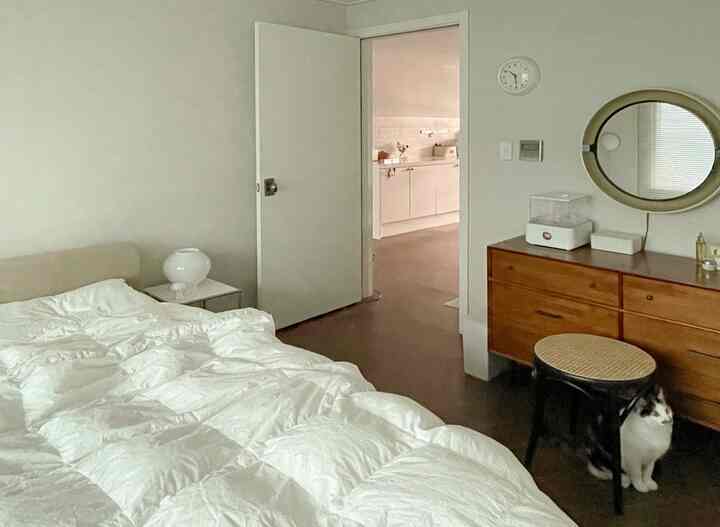 White and natural brown toned bedroom featuring wooden dresser, round mirror, and a cat sitting beside a stool, creating a cozy atmosphere
