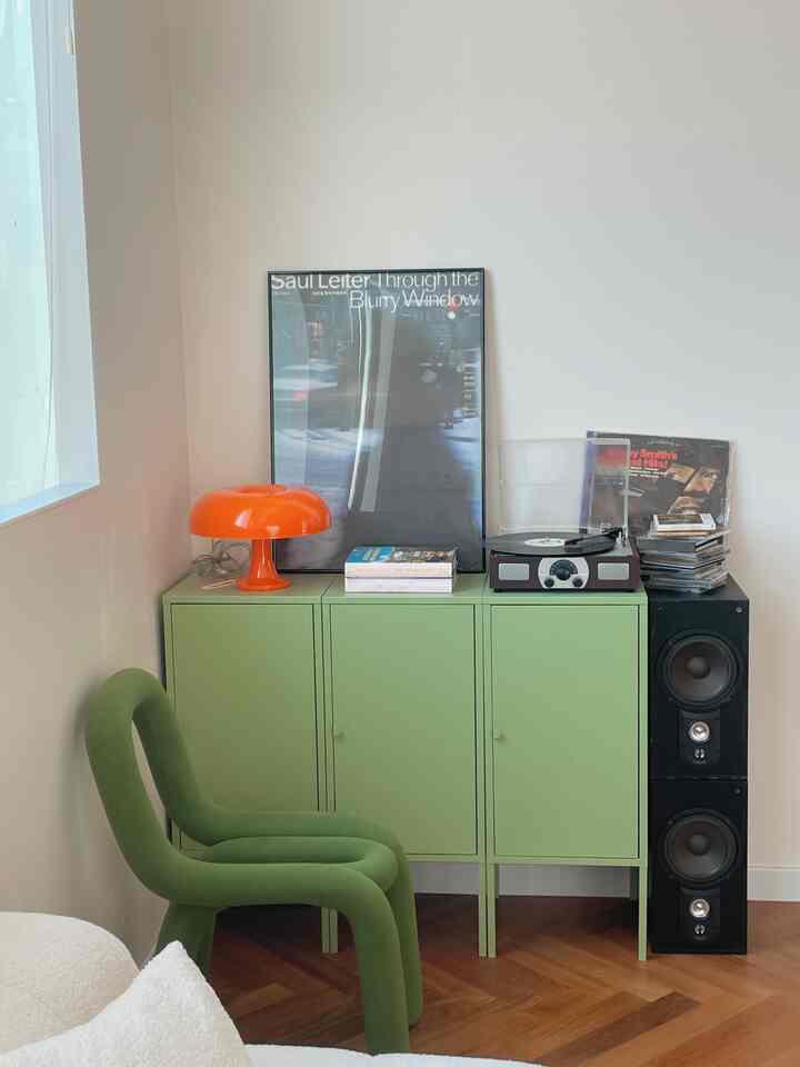 A retro-style living corner with green cabinet and chair, orange table lamp, and wooden floor against light cream walls
