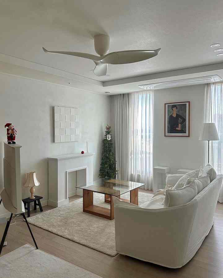 明るいホワイトトーンのリビング、ホワイトファブリックソファとガラスのテーブル、ラグ、暖炉コンソール、シーリングファンが配されたミニマルなインテリア