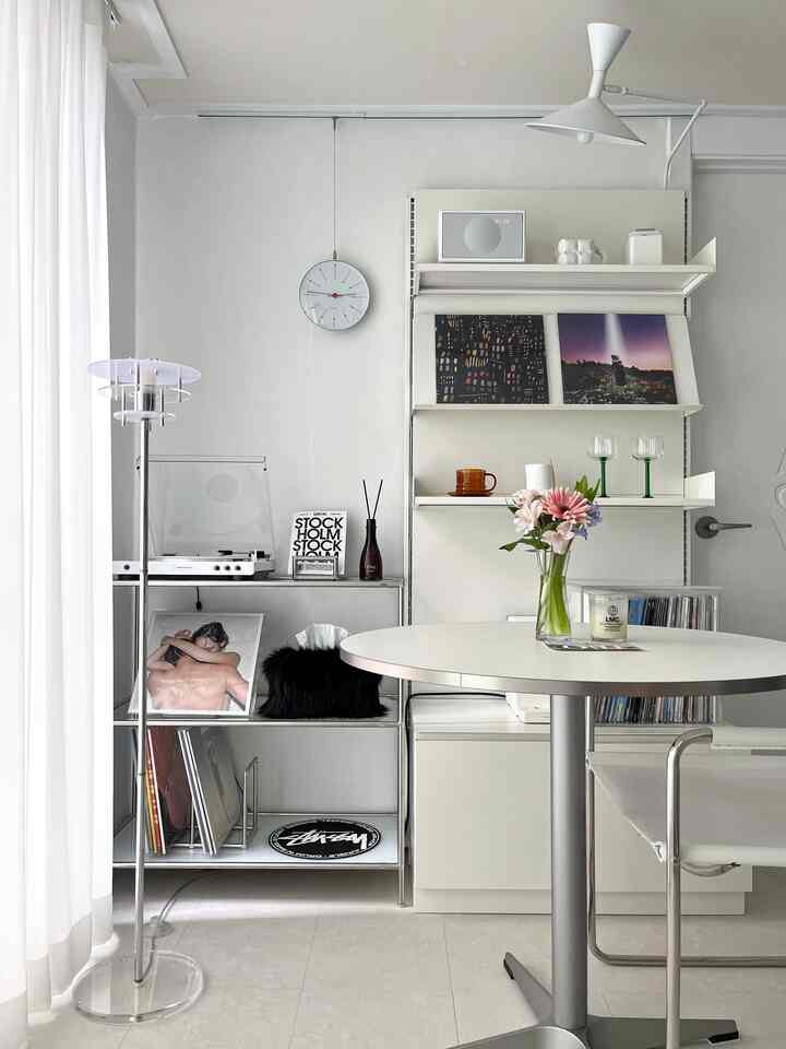 Modern living room in white and light gray tones featuring a round table, shelves, and bright sheer curtains