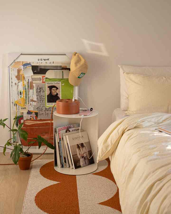 Natural-toned bedroom with brown wooden floor and white walls, beige bedding, featuring side table, picture frame, and plant creating a simple, calm atmosphere