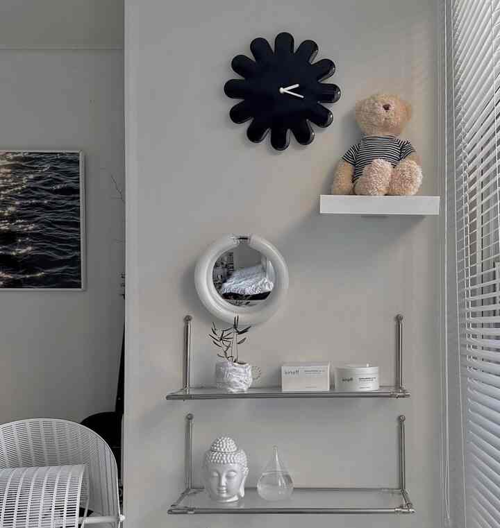A simple living room wall in white and modern gray tones featuring a black wall clock, shelves with a teddy bear, and decorative items