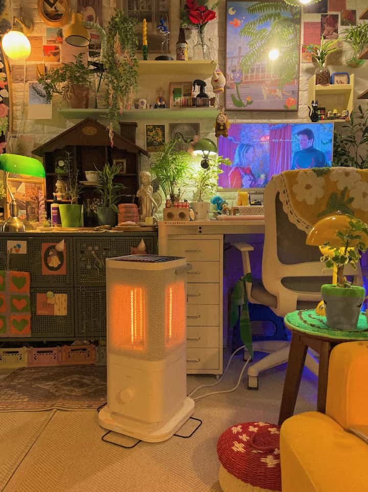 暖かいイエローとグリーンの色調のリビング、オバンストーブの電気ヒーターと植物、ヴィンテージ家具が配置された雰囲気のあるインテリア