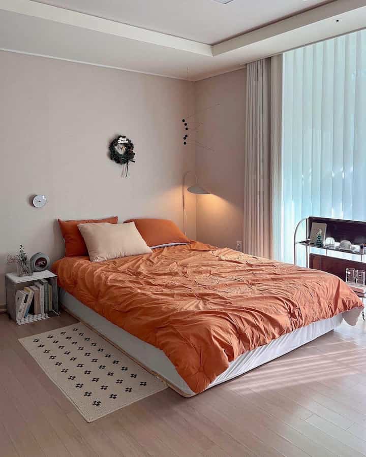 ベージュの壁とカーテンが調和した居心地の良い寝室、オレンジの寝具と壁の照明が特徴のナチュラルモダンなインテリア