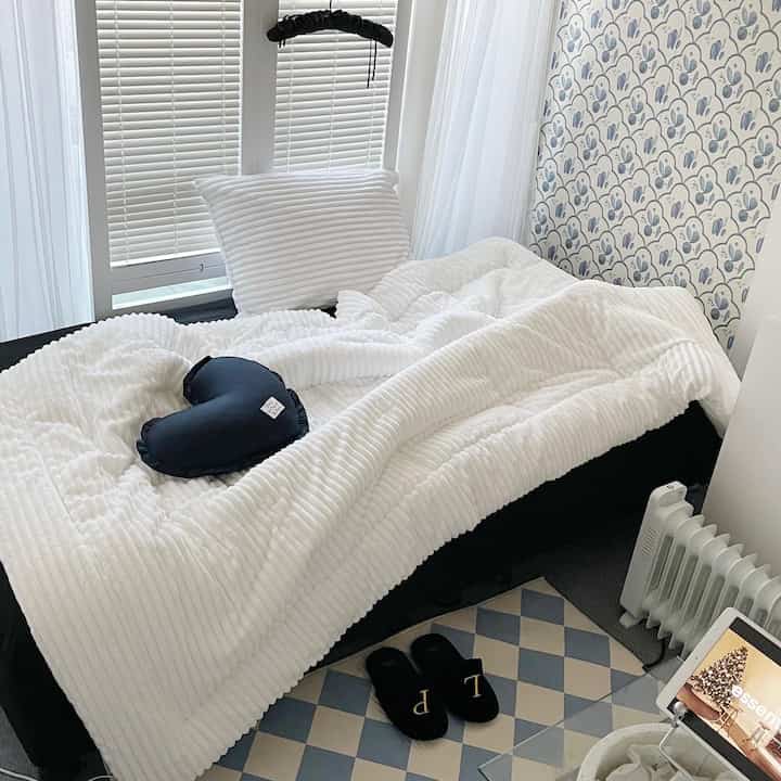 Small white-toned bedroom featuring fluffy microfiber bedding and an electric heater, creating a cozy atmosphere