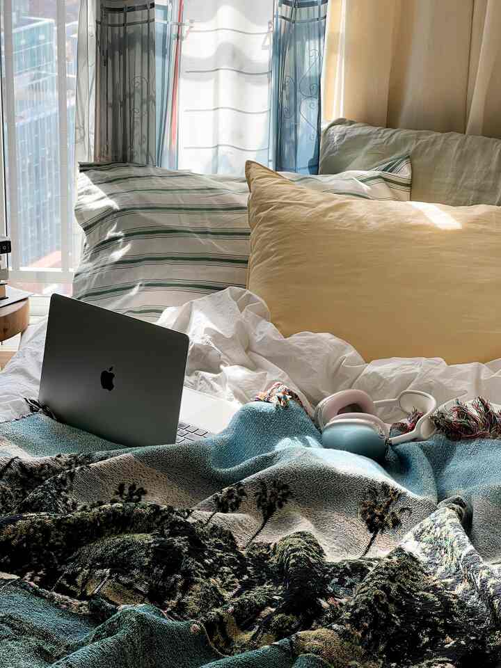 Natural-toned bedroom featuring white bedding, pillows, curtains by the window, and a laptop creating a cozy atmosphere