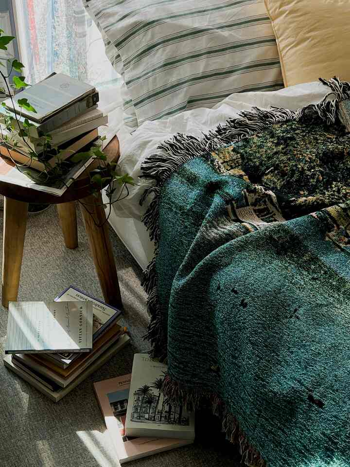 Natural color and wood tone bedroom featuring a bed with pillows and blanket, books stacked on a wooden stool creating a cozy reading nook