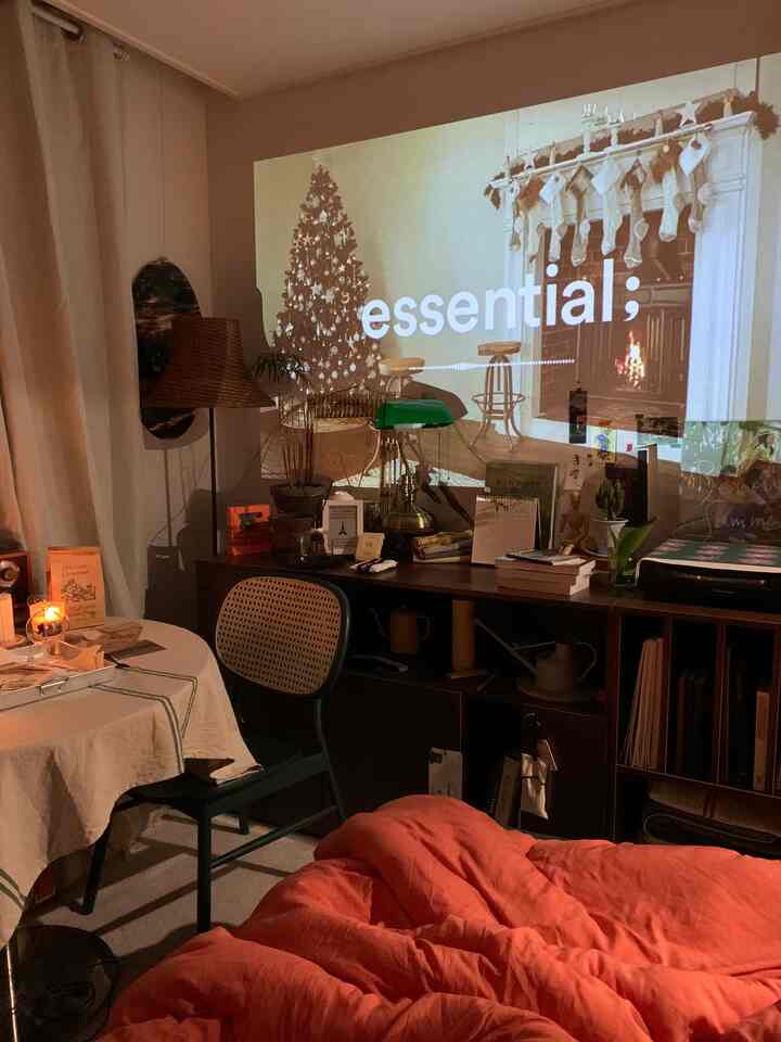 暖かみのある木目調とオレンジの寝具が調和するリビング、壁にクリスマスと暖炉の映像が映し出された雰囲気のある空間