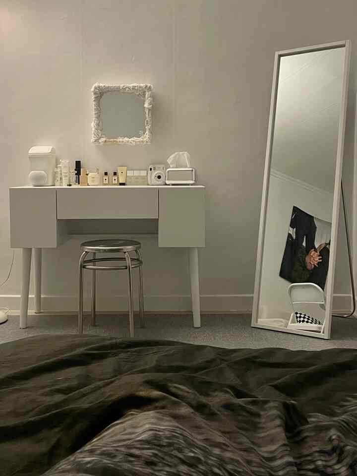 Bright white-toned bedroom vanity area featuring a vanity table with stool at center and a full-length mirror leaning on the right wall, simple and calm interior