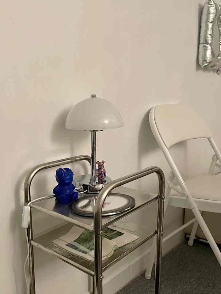 Simple space with white walls and folding chair, silver metal side table topped with table lamp and miniature figures