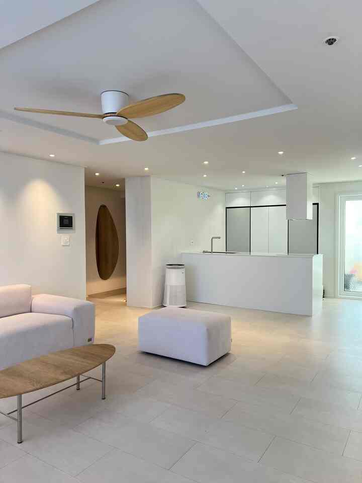 A white and light brown toned studio apartment living room and kitchen space featuring modern sofa, coffee table, and ceiling fan in a clean interior