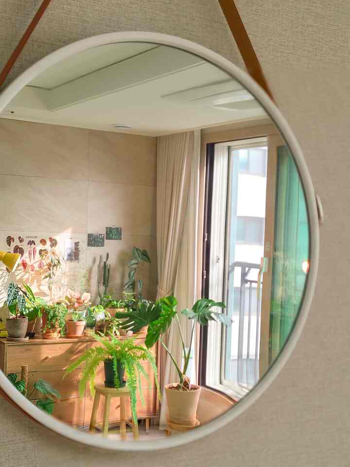 Natural wood tones and ivory curtains blend with plants reflected in a round mirror creating a cozy balcony interior space