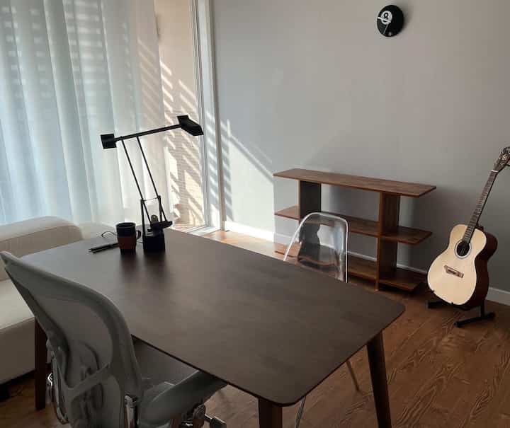 Bright modern living room with wood tone flooring and simple wooden dining table, featuring transparent chairs and sheer curtains controlling natural light