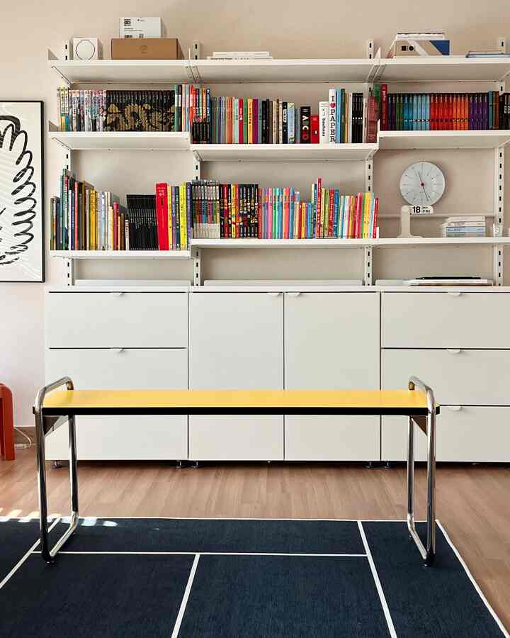 白を基調とした壁と本棚、ネイビーのラグに黄色いベンチが配置された実用的なリビングスペース