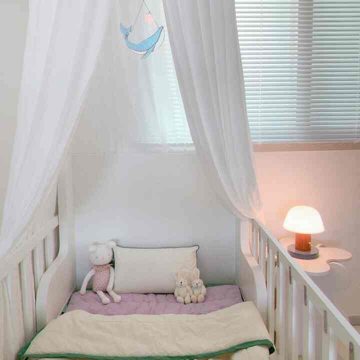 White and lavender toned kids' room featuring a white bed, sheer curtains, and a soft table lamp creating a cozy baby bedroom