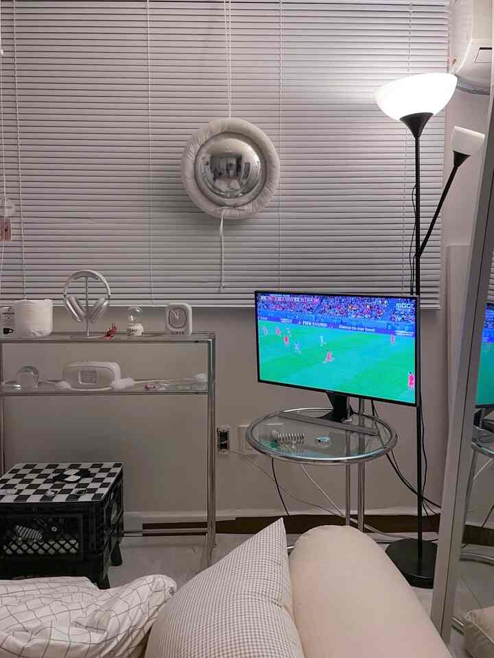 白とグレーのトーンのリビングルーム、ブラインドの窓とフロアランプ、モダンなサイドテーブルとクッションが配置された落ち着いた空間