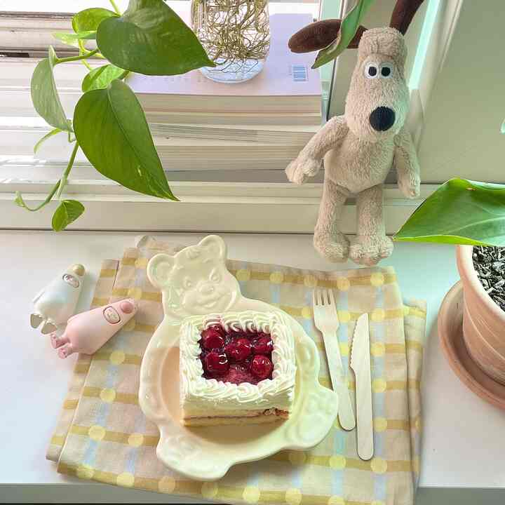 温かみのあるベージュとグリーン色調の窓際のテーブル、クマ形皿や植物、犬のぬいぐるみが印象的な空間