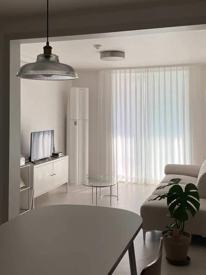 明るい白色調のリビング、ミニマルな白いソファと透明なガラスのコーヒーテーブル、大きなミルクシフォンカーテンが配置された清潔感のあるインテリア
