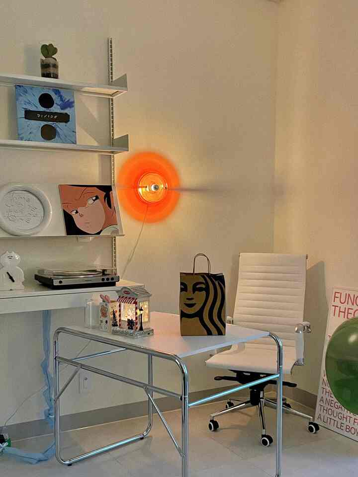 A simple white home office with an orange wall sconce, featuring a desk, an office chair, and organized shelving