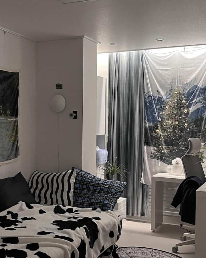 白と黒を基調としたミニマルな寝室、デスクや椅子、カーテン、クリスマスツリーのファブリックポスターが配置された落ち着いた空間