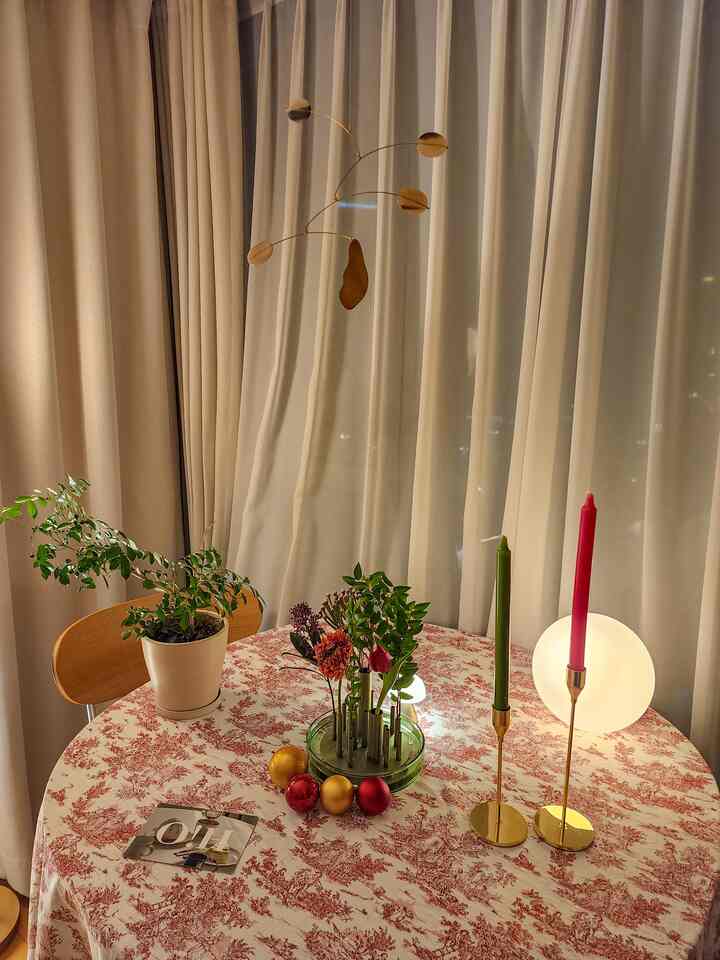 Beige and ivory curtains frame a dining table featuring a vintage patterned tablecloth, gold candle holders, a vase, and plants in a natural setting