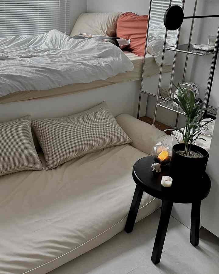 A cozy studio bedroom with white and natural tones, featuring a large floor cushion and a black side table