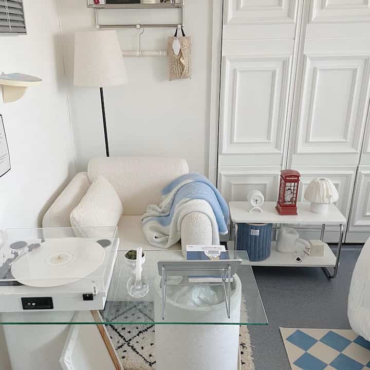 White and blue toned studio apartment living room featuring a single-seat sofa, glass desk, and turntable in a clean modern space
