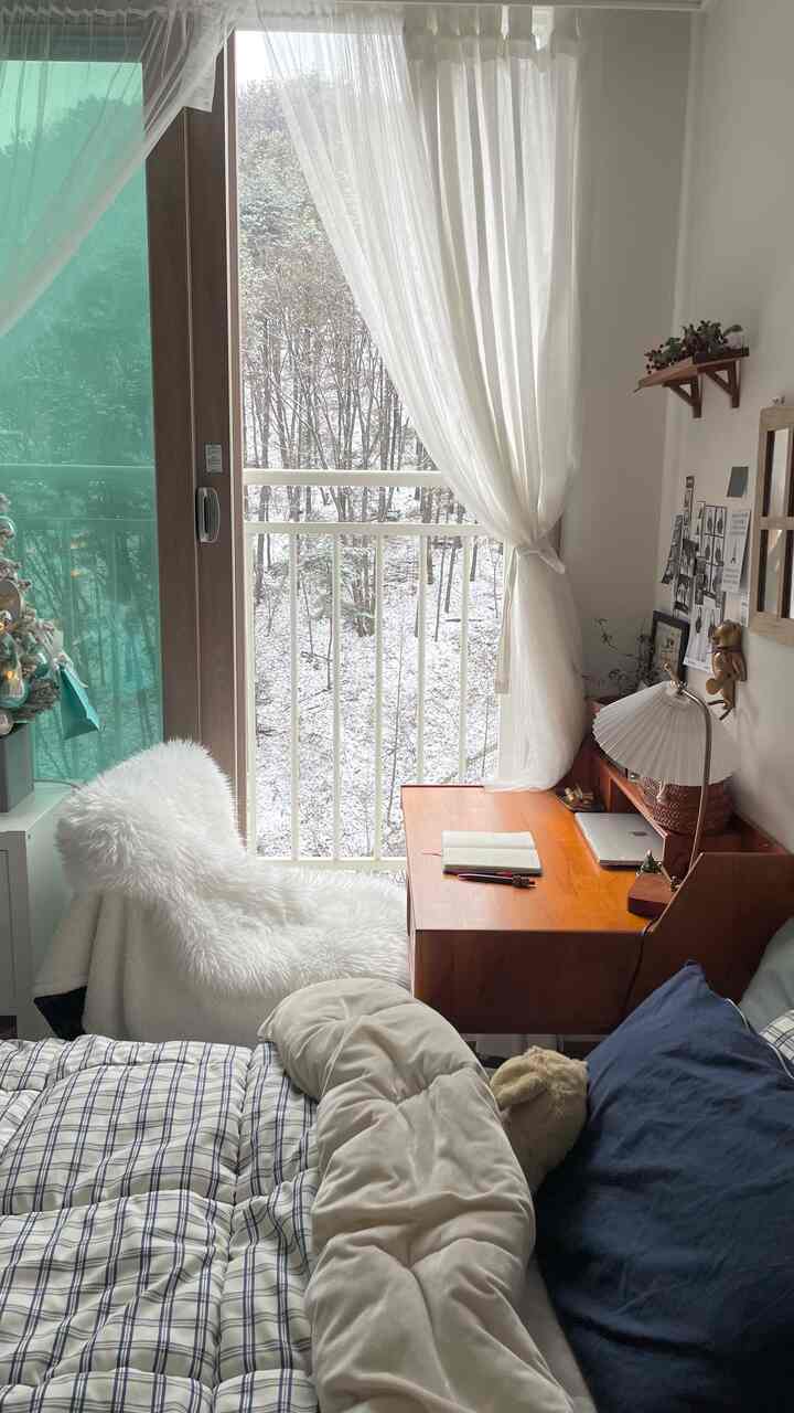 A white-curtained compact bedroom with natural wood desk set up as home office for a single household, snowy outdoor view through window