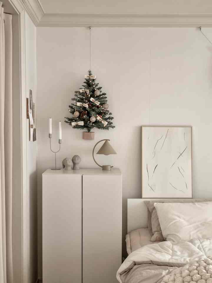 ホワイトとブラウンのトーンの寝室、壁に掛かった小さなクリスマスツリーとシンプルなキャビネットが配置された落ち着いた雰囲気の空間