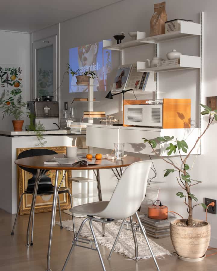 Warm natural light fills a Nordic-style kitchen-dining space featuring a wooden dining table, shelves, and various plants for a cozy atmosphere