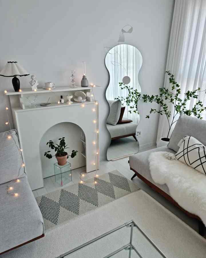 A white and gray toned living room featuring an irregular full-length mirror, fireplace console, and plants in a simple modern setting