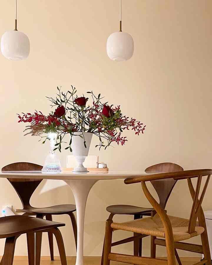 A round dining room with soft beige walls and wooden dining chairs, featuring a white vase with red flowers on a round white table at the center