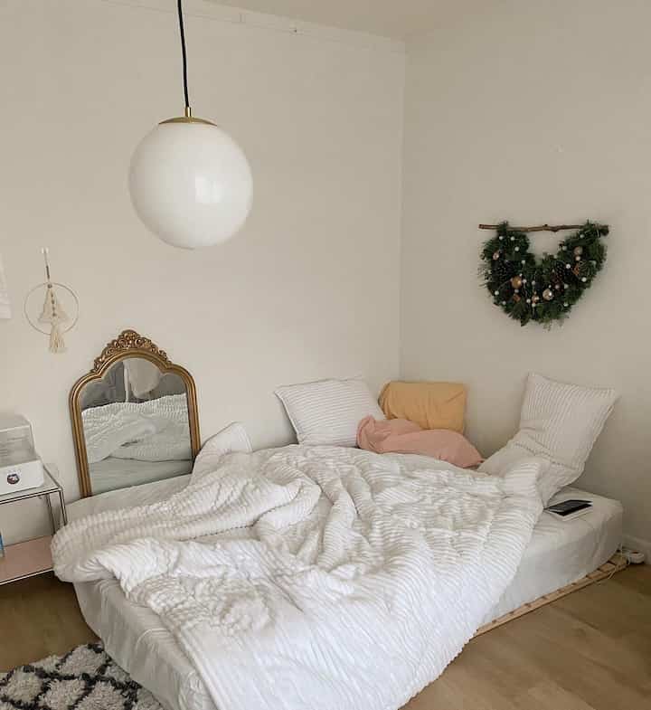 A cozy bedroom with white and natural wood tones, featuring a large pendant light, a gold ornate mirror, and a Christmas wreath on the wall