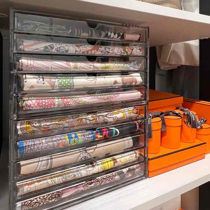 透明なアクリル引き出しに整理されたカラフルなスカーフと鮮やかなオレンジ色の高級パッケージが並ぶナチュラルトーンのウォークインクローゼット