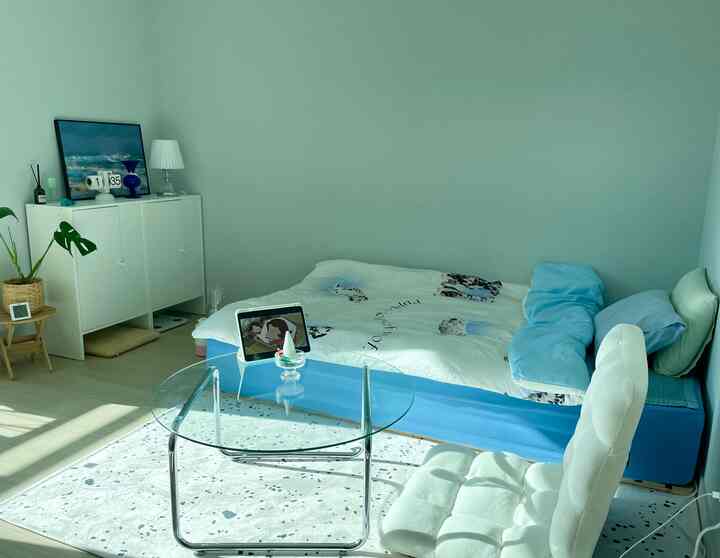 Pastel-toned single bedroom featuring a blue bed, white cushioned chair, and a transparent round glass table in a clean, compact space