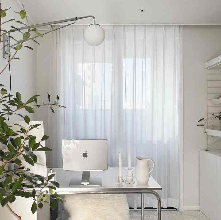 White-toned study room featuring a central desk with laptop, candles and pitcher nearby, and a green plant on the left, creating a clean atmosphere