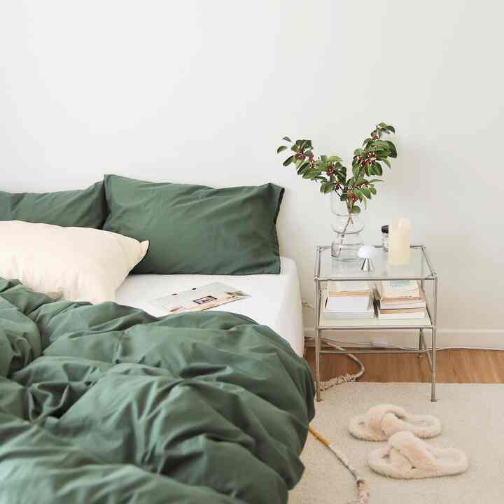 白い壁と木の床の寝室、緑の寝具とクッションのベッド、ガラスのナイトテーブルが落ち着いた雰囲気を演出