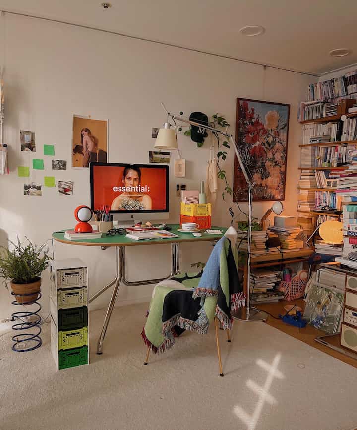 Bright cream walls and rug define a mid-century modern home office featuring a green desk, bookshelf, and plants for a cozy atmosphere