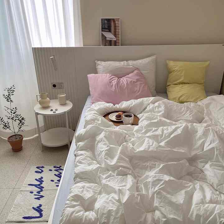 A bedroom featuring white and pastel tones, with white bedding, pink and yellow pillows, a round nightstand, and a small plant creating a cozy atmosphere.