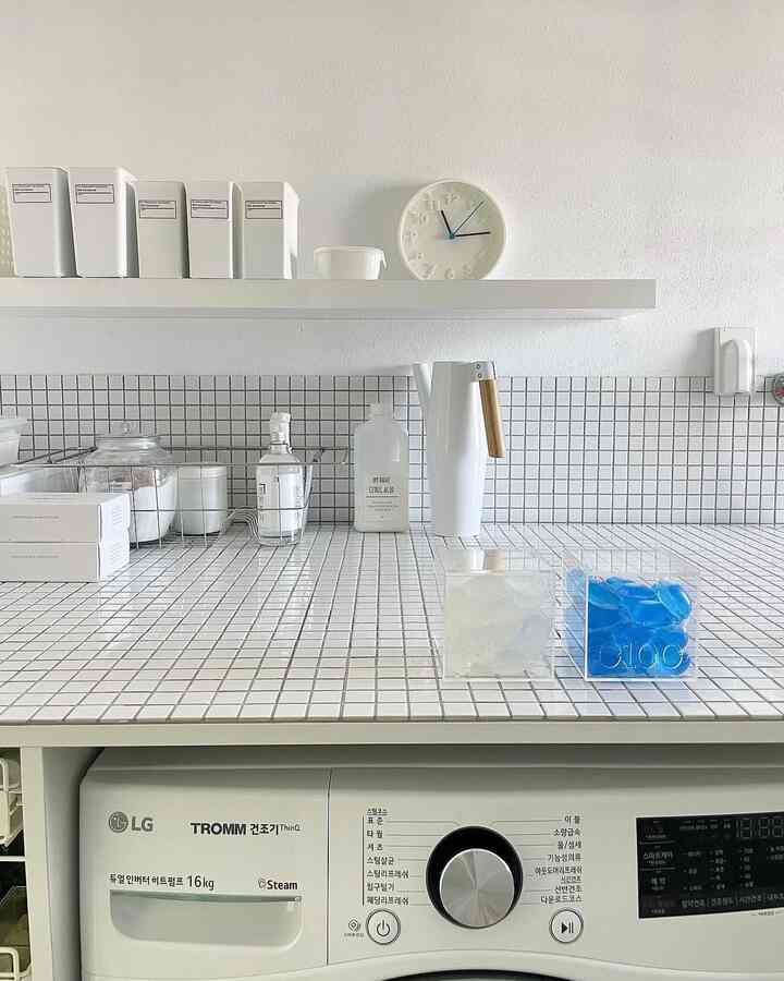 Compact laundry room with white tiled countertop and shelf, featuring organized storage containers, washing machine, dryer, and a clock, creating a clean interior